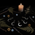 Five silver crescent moon stitch markers arranged in an arc on black velvet with moonstones and candle bokeh