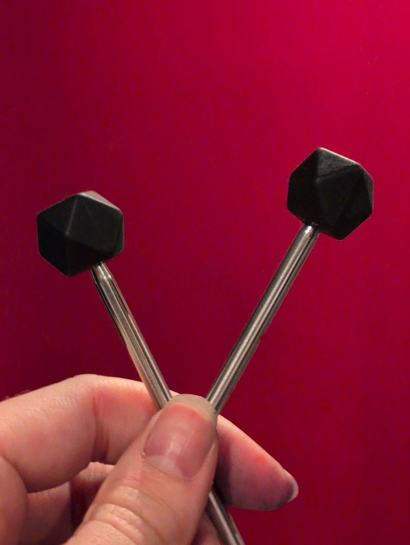 A close up on a hand holding two knitting needles with black silicone hexagonal stitch markers on them.